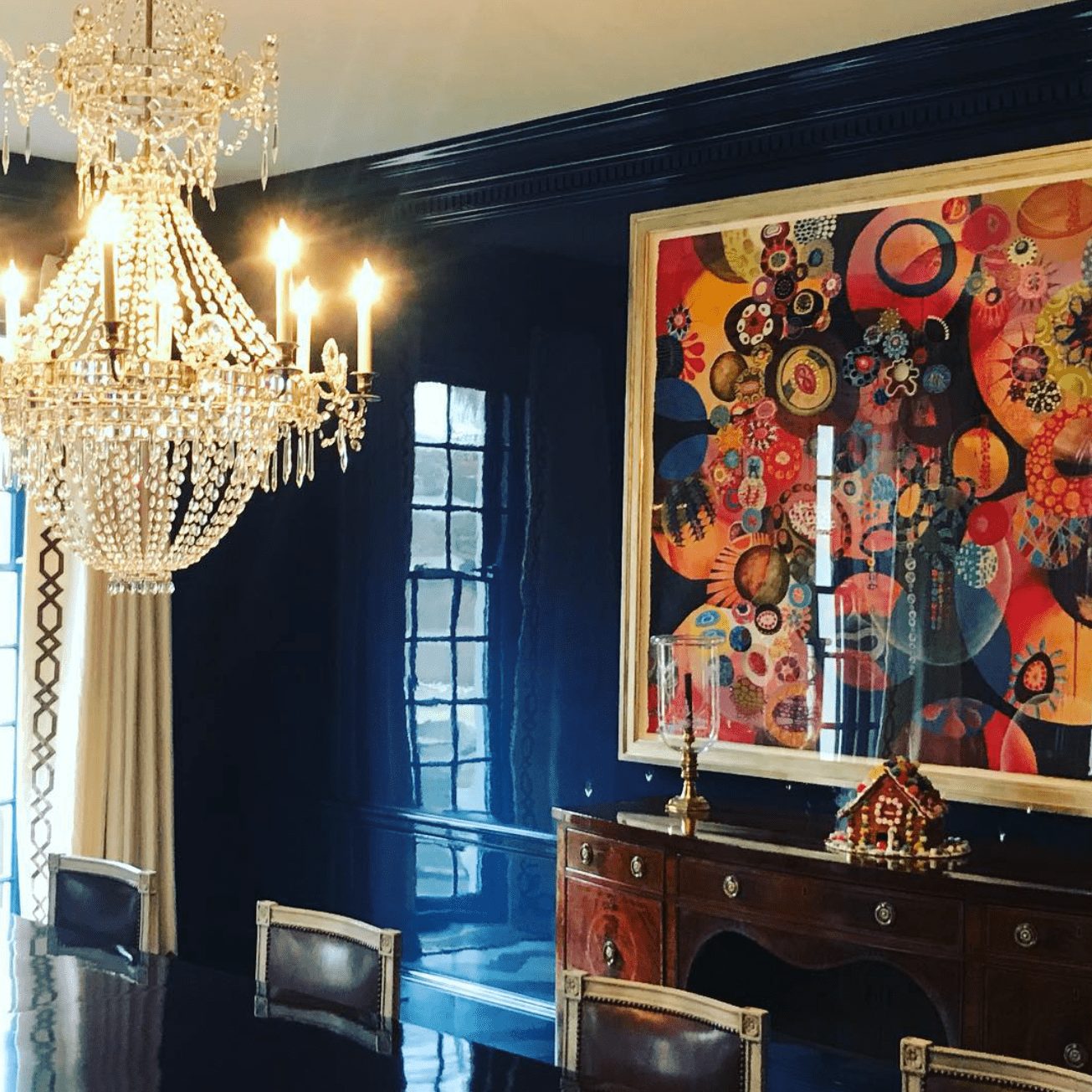 A glossy black accent wall in a dining room. There's a colorful painting and an expensive chandelier around a shiny dining table. 